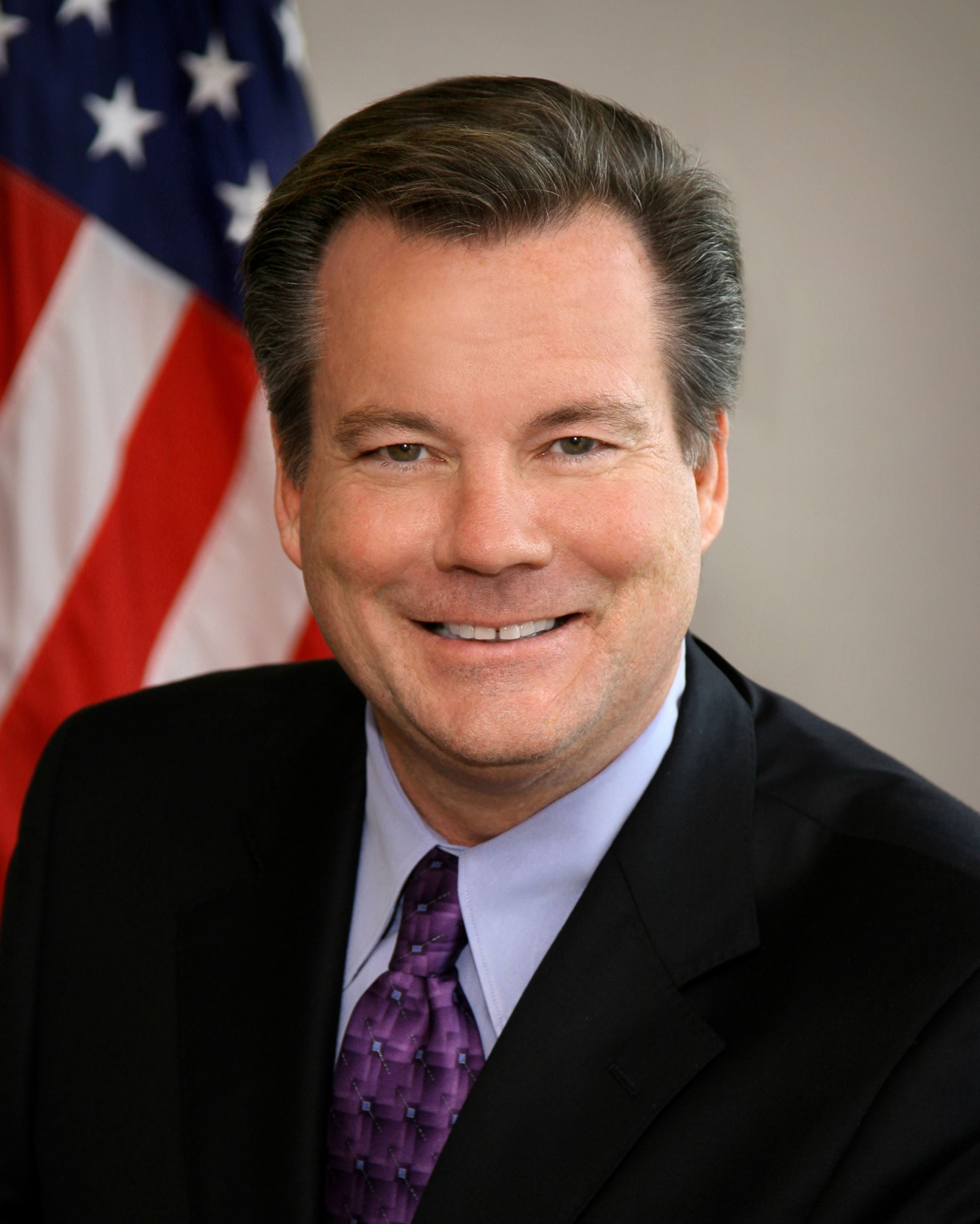 Commissioner Tom Shelly smiling in front of an American flag.