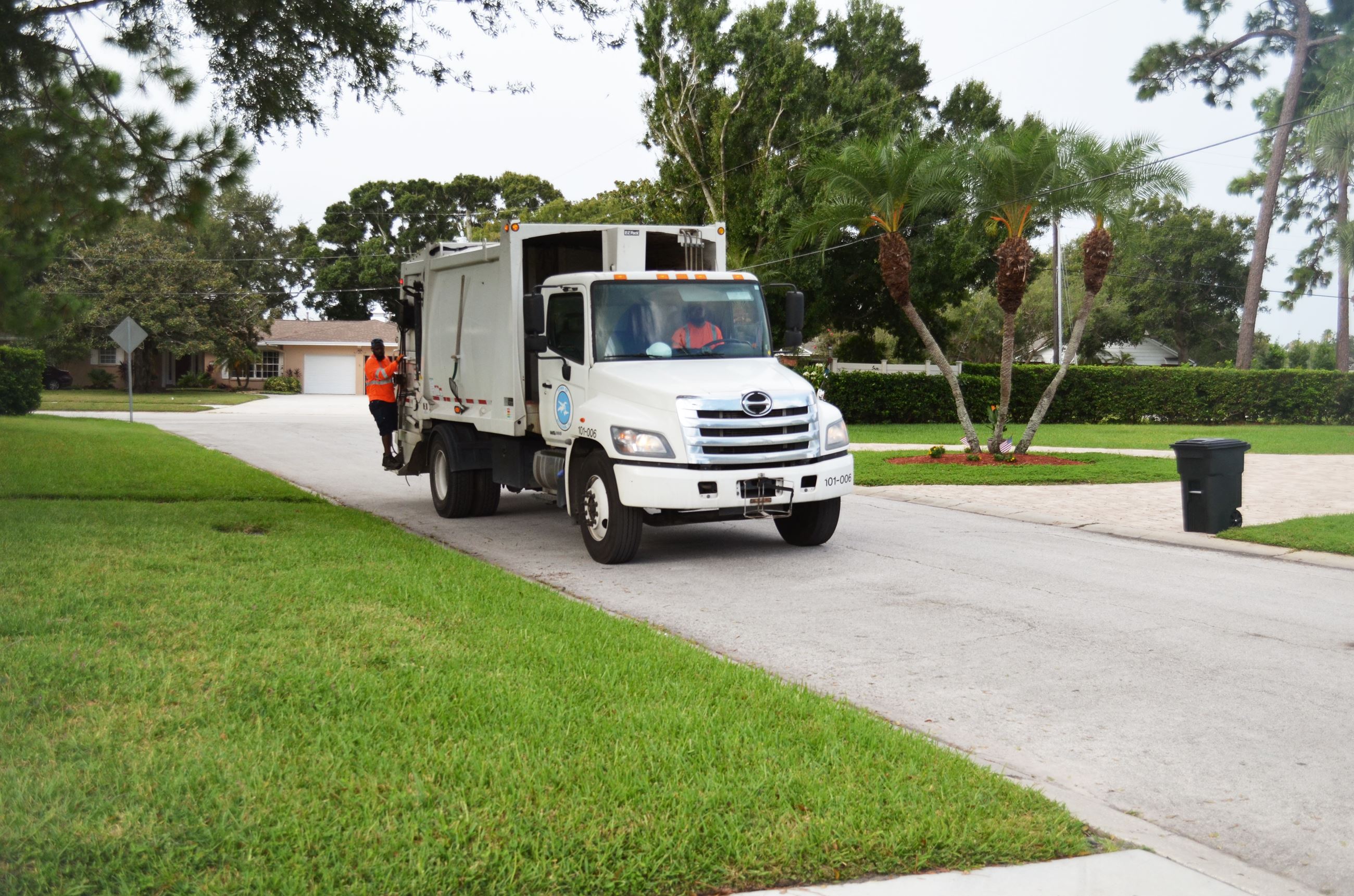 A solid waste employee rides on the back of a garbage truck towards a full can.