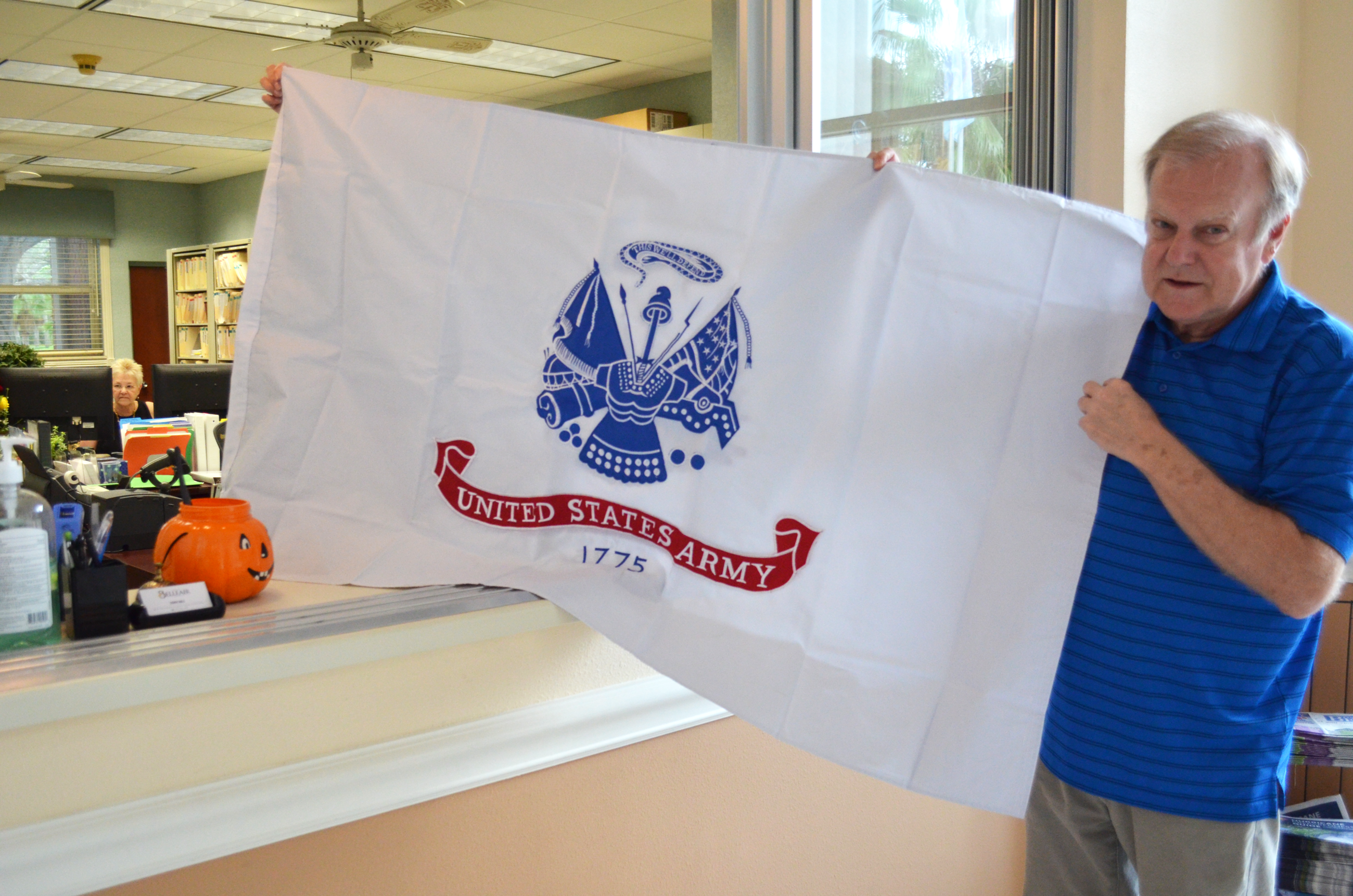A man holds a US Army flag in the front lobby of Belleair Town Hall. Below, a caption 