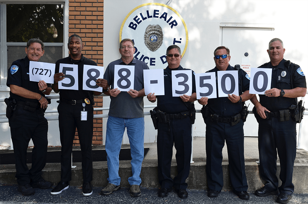 Six Belleair Police Officers stand outside of the police building holding sheets of paper with their